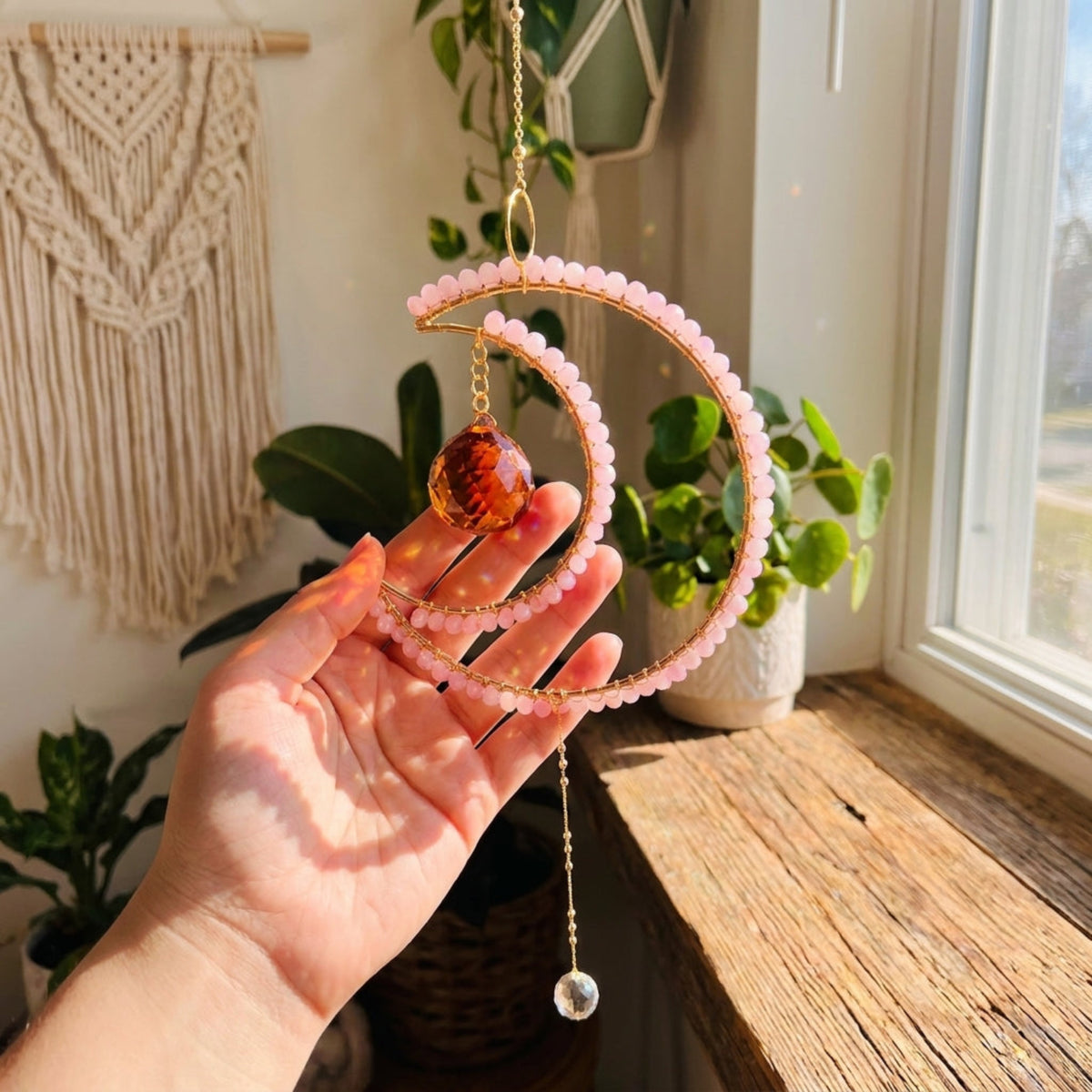A hand holds a gold crescent moon suncatcher ornament with pink beads and a brown amber glass orb, set against a warm, sunlit room background with plants and a macrame wall hanging.
