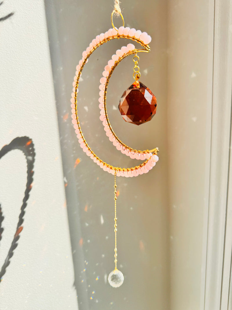 Decorative hanging moon with pink beads and a red gemstone against a neutral background