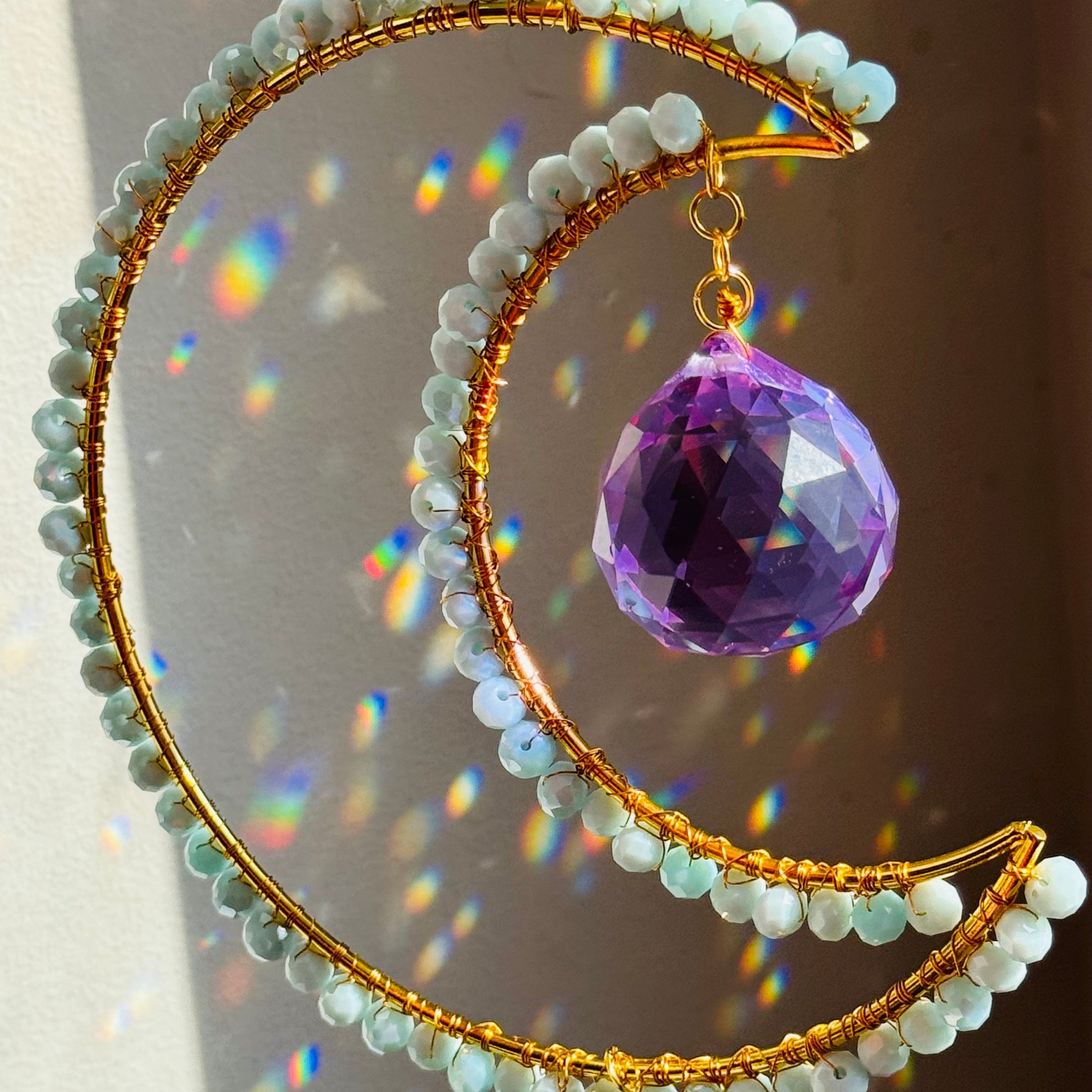 A crescent-shaped suncatcher with a golden frame, adorned with a variety of blue and clear beads, and a large purple bead in the center.