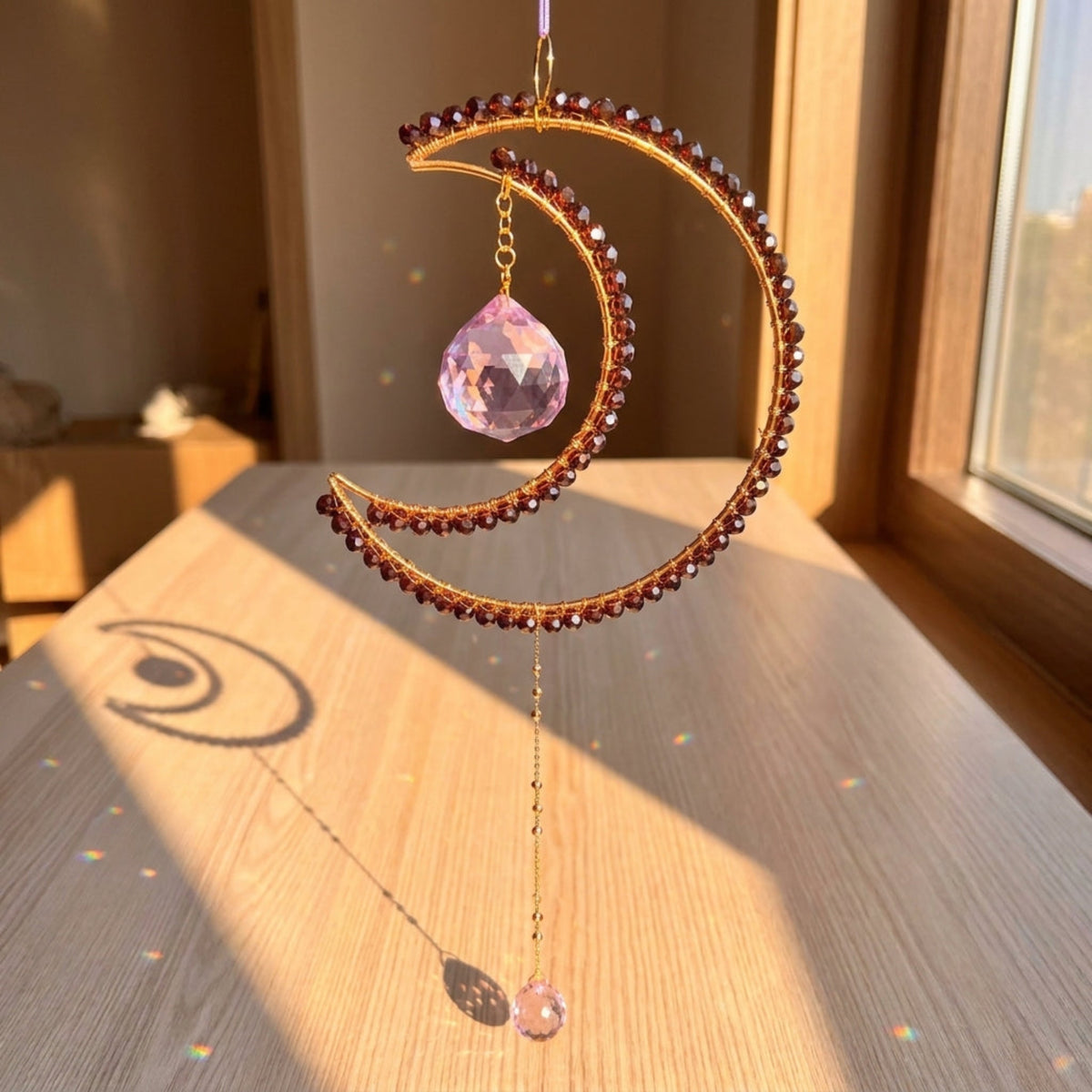 A crescent moon-shaped gold-toned beaded wall hanging ornament with two faceted light purple crystal drops, hanging and creating rainbows near the window and a table.