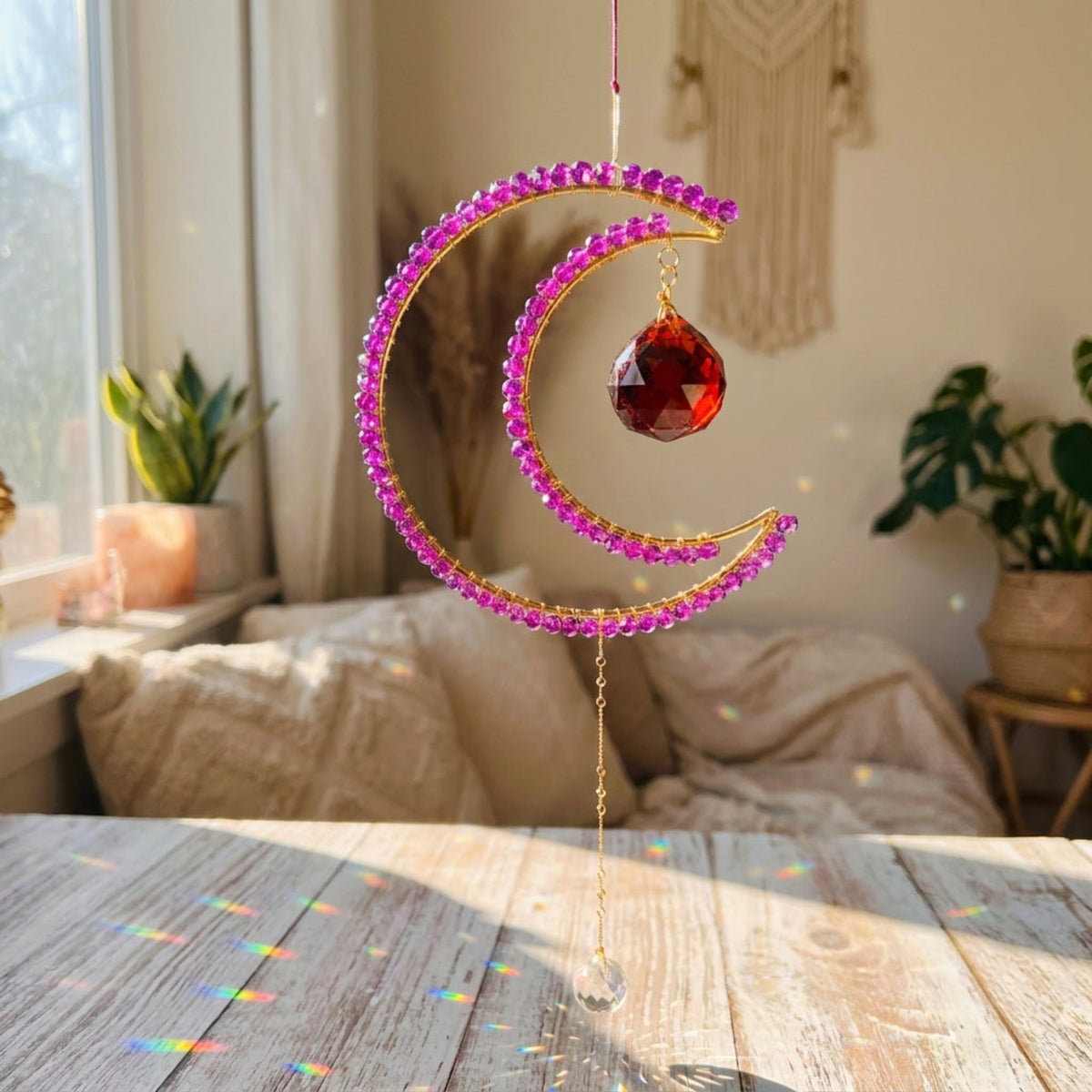 A gold crescent moon suncatcher ornament adorned with pink rhinestones and a faceted red crystal, hanging on a wooden table in a sunlit bedroom with plants and a bed in the background.