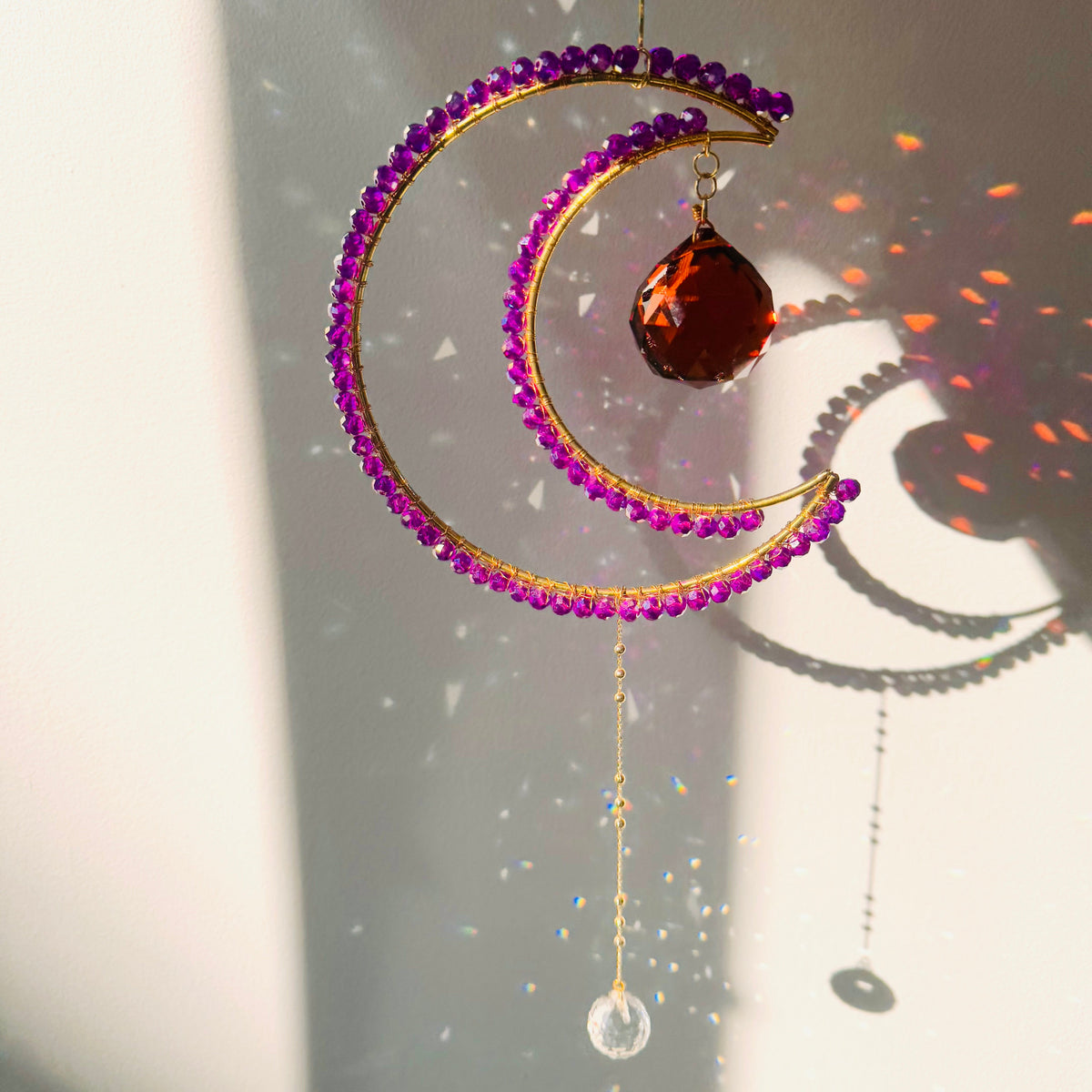 A crescent-shaped suncatcher with magenta colored beads and a crystal pendant, hanging by a string against a sunlit background.
