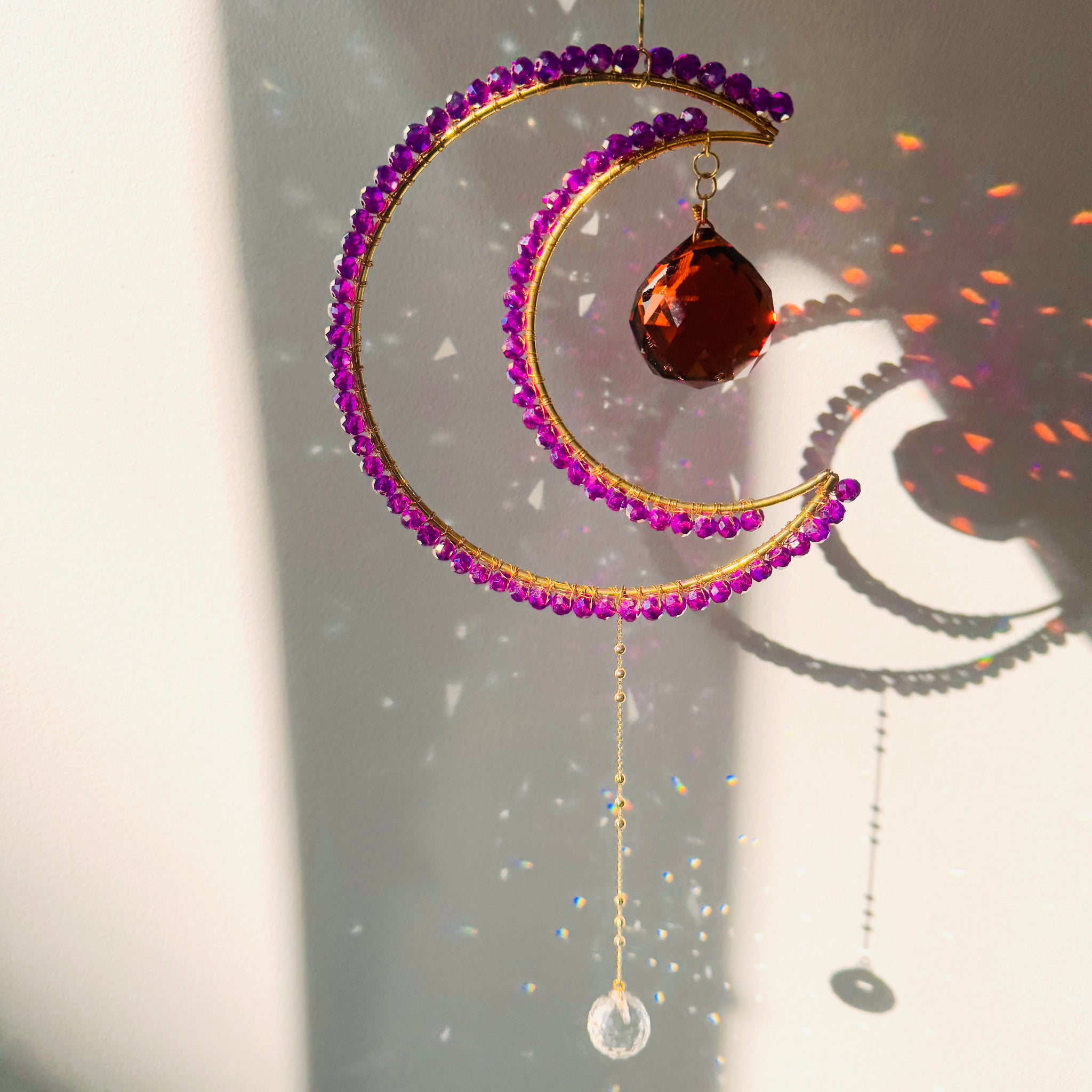 A crescent-shaped suncatcher with magenta colored beads and a crystal pendant, hanging by a string against a sunlit background.