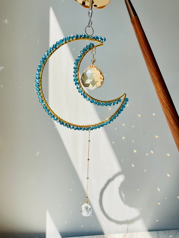 A crescent-shaped suncatcher with blue beads and a crystal pendant, hanging by a chain, with light reflecting off the beads.