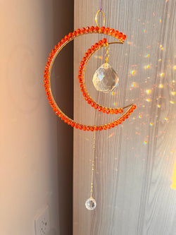 A crescent-shaped orange suncatcher with wire-wrapped beads and a crystal prism hanging against a window with light shining through, creating a colorful refraction.