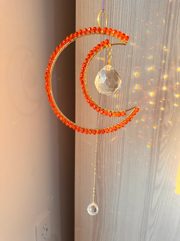 A crescent-shaped orange suncatcher with wire-wrapped beads and a crystal prism hanging against a window with light shining through, creating a colorful refraction.