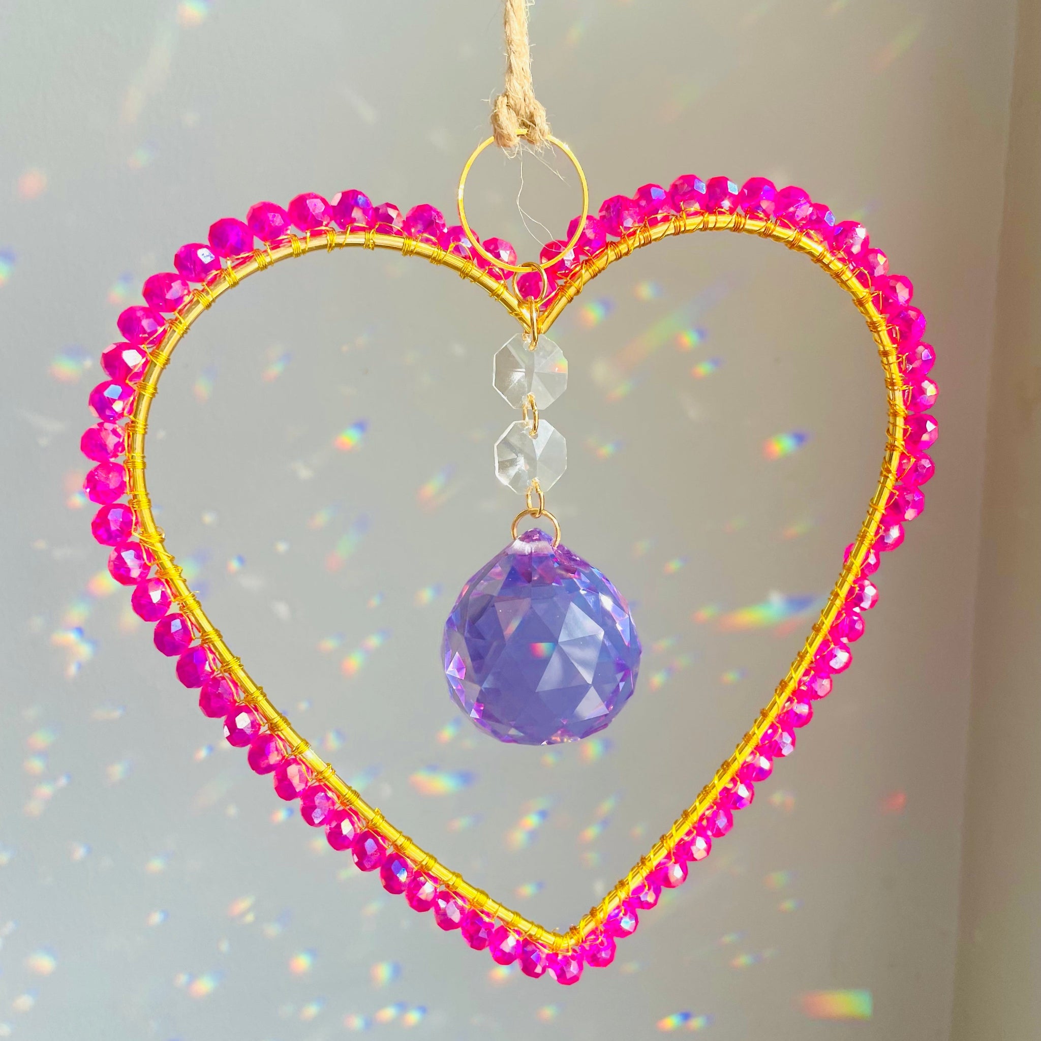 A handcrafted magenta crystal suncatcher with a heart shape, hanging by a string, with a large lavender prism at the center surrounded by smaller pink crystals.