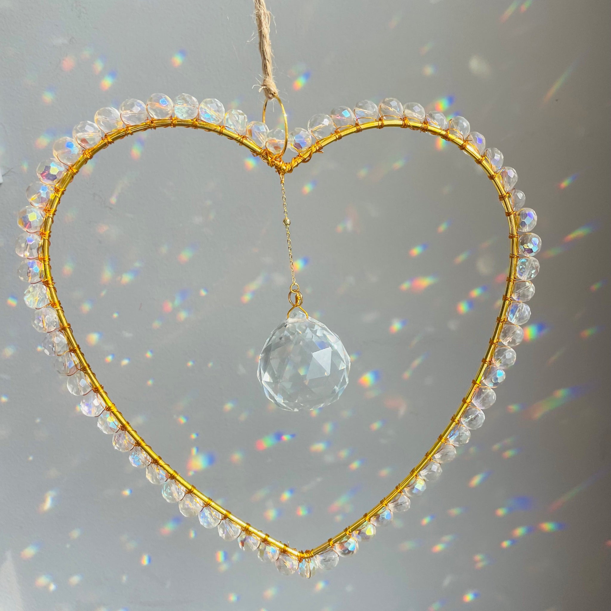 A heart-shaped clear suncatcher with gold-colored wire wrapping and a crystal prism, hanging against a reflective background.