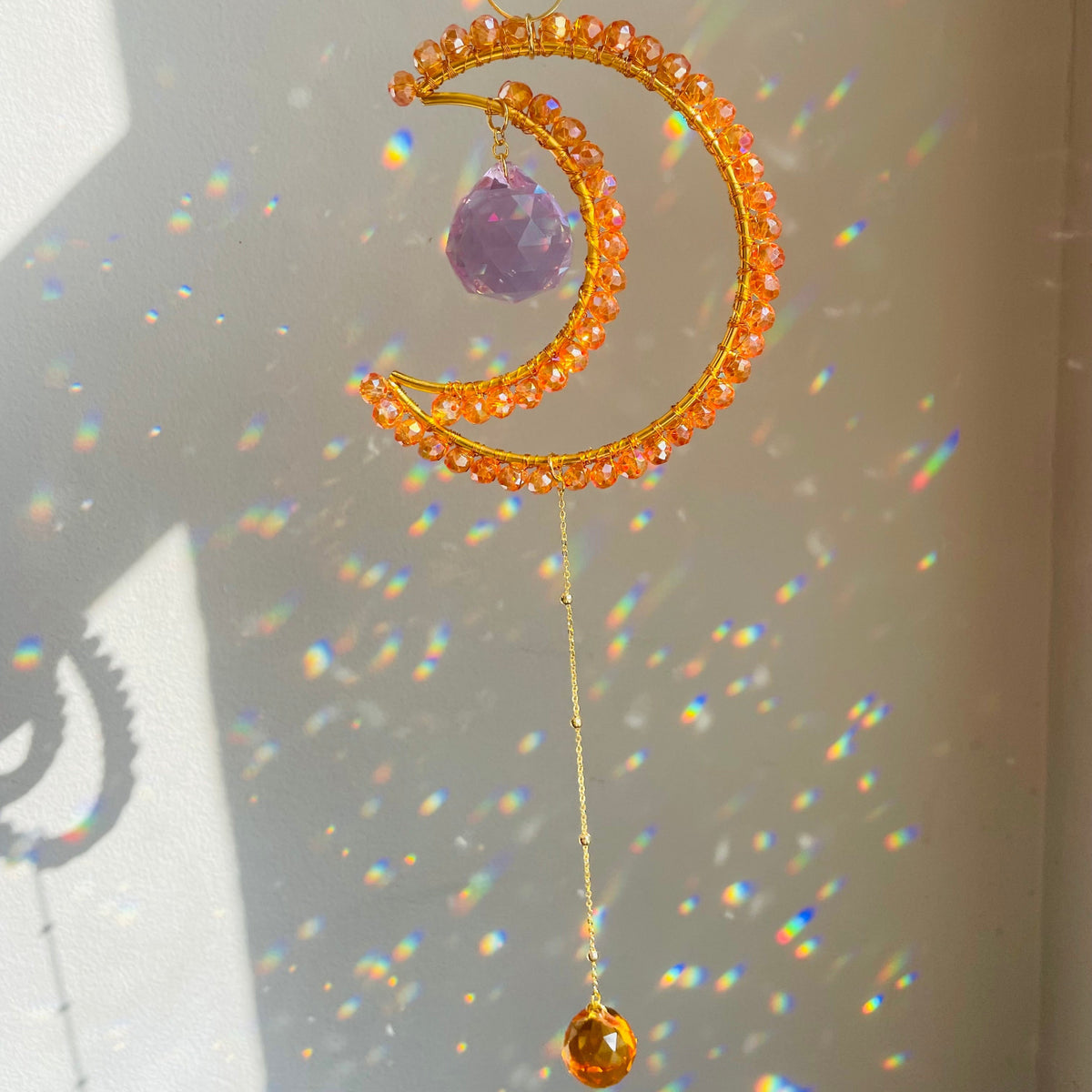 A crescent-shaped orange suncatcher with a purple center and a yellow hanging loop, displayed against a backdrop of a sunlit wall.