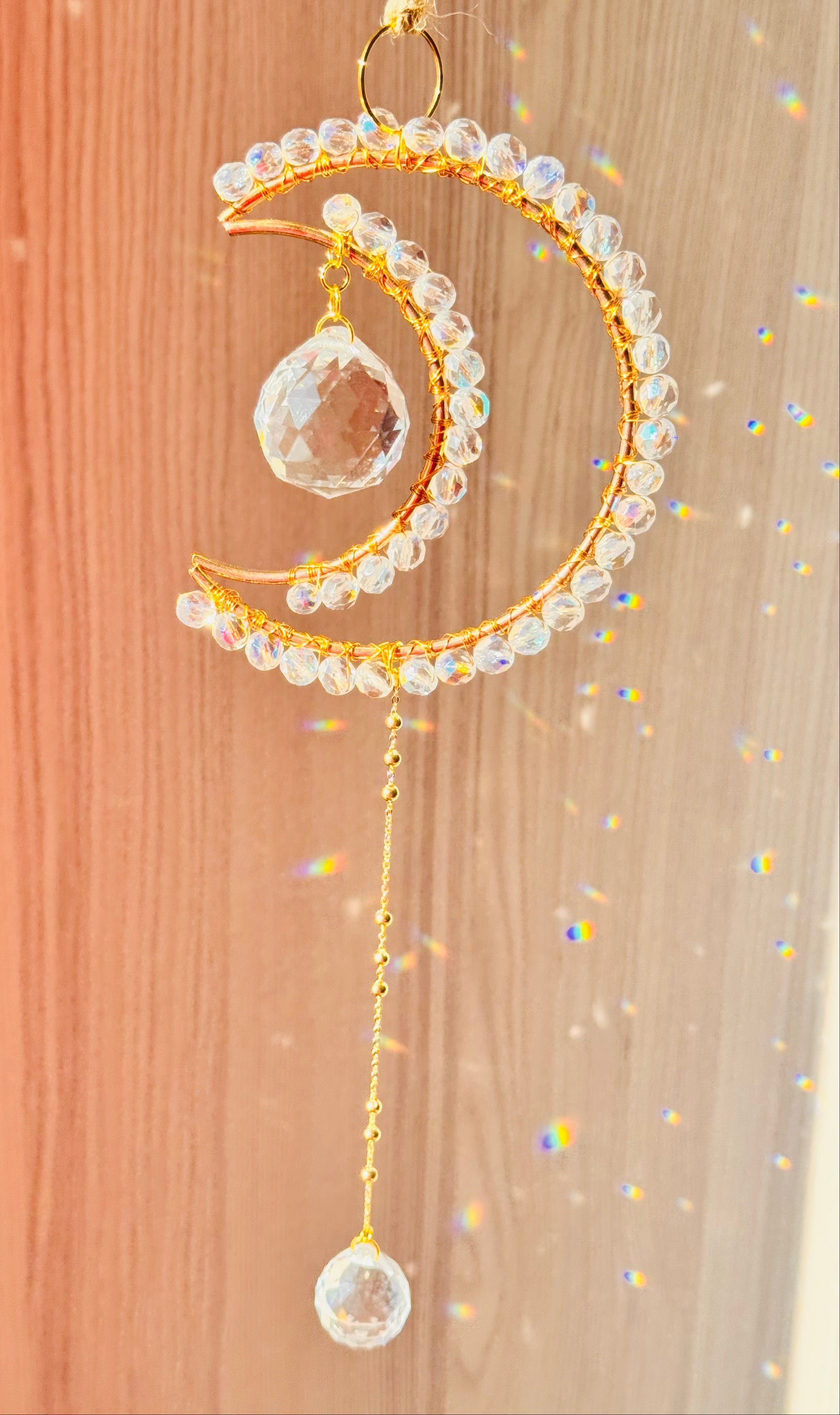 A gold-colored crescent-shaped suncatcher adorned with clear crystals and a large clear prism at the bottom, hanging against a pink backdrop.
