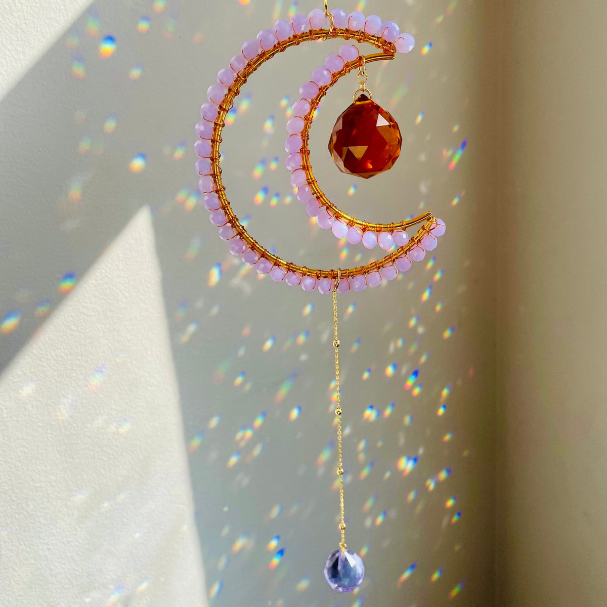 A crescent-shaped suncatcher with a lavender crystal in the center, dangling from a chain of beads, with a play of light reflections on the wall.