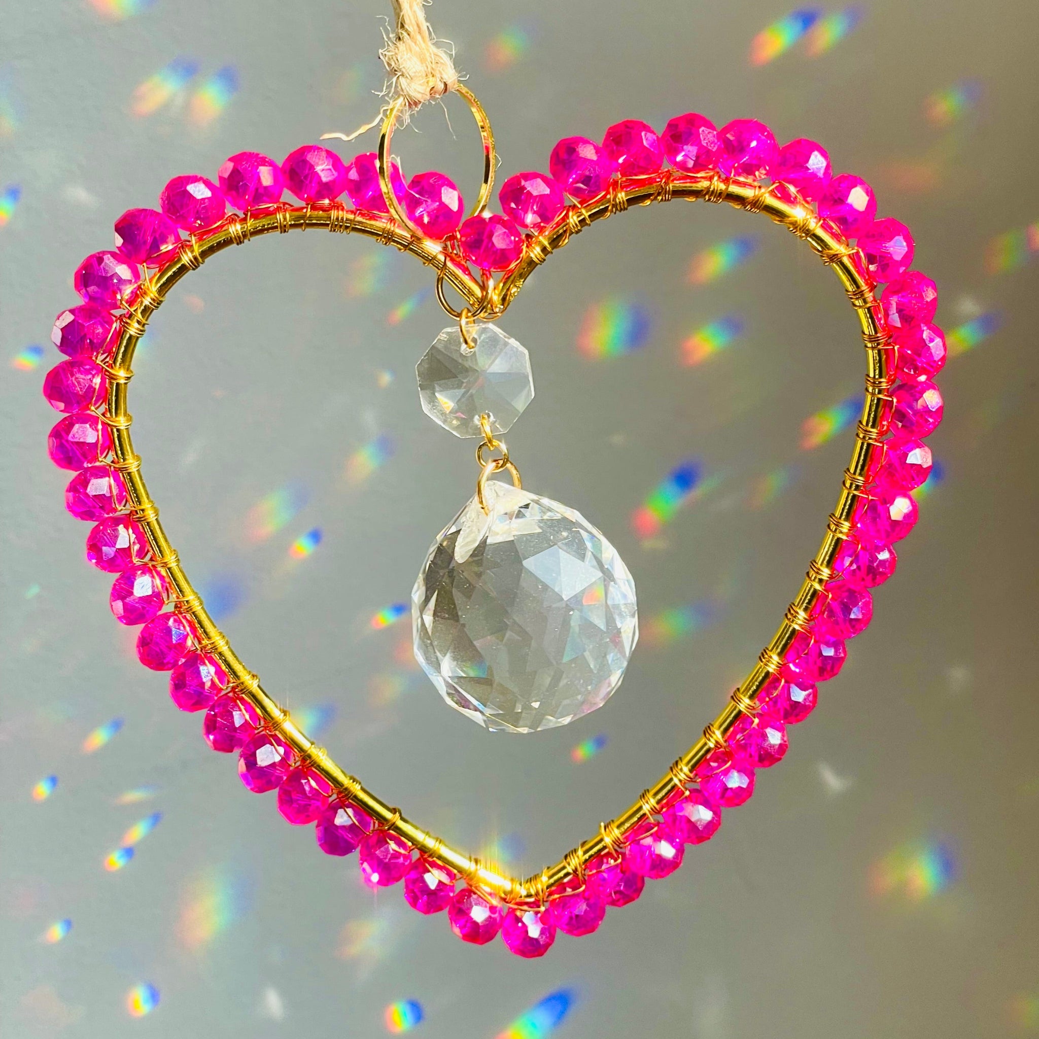 A gold-colored heart-shaped suncatcher with wire-wrapped Czech crystals arranged around a faceted glass prism, hanging by a string against a plain background.