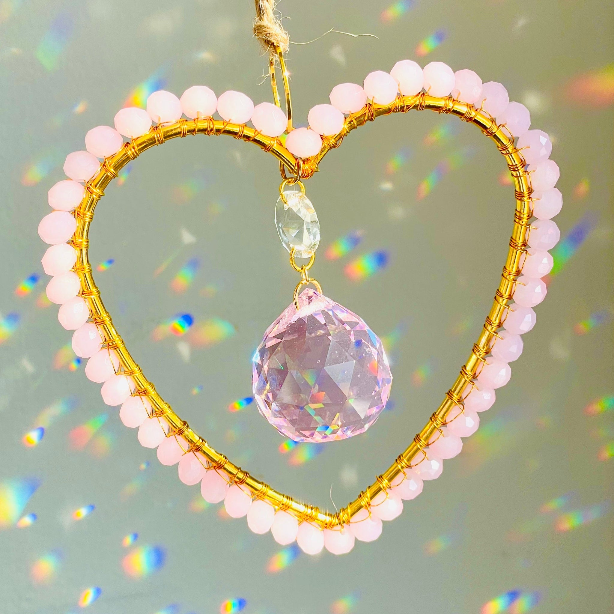 A gold-colored heart-shaped suncatcher with wire-wrapped Czech crystals arranged around a faceted glass prism, hanging by a string against a plain background.
