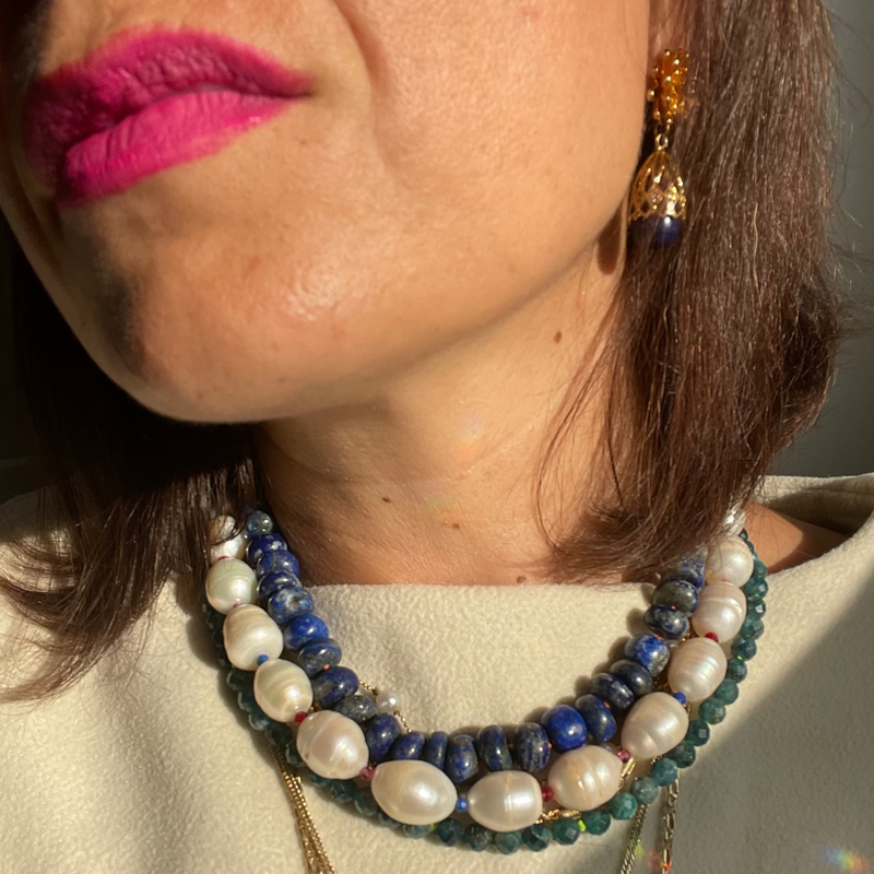 Close-up of a person wearing multiple necklaces with beads and a gold chain, with a neutral background.