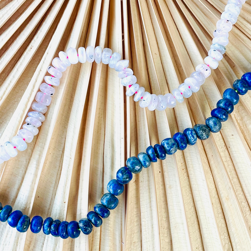 Two beaded necklaces, one white and one blue, on a textured beige surface.