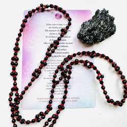 A black obsidian pendant with a red and clear beaded necklace, placed on a white surface with a purple text card that contains poetry.