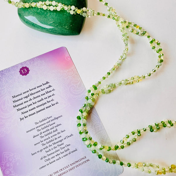 A tantric necklace with green gemstones and a green heart-shaped bead, accompanied by a card with spiritual text.