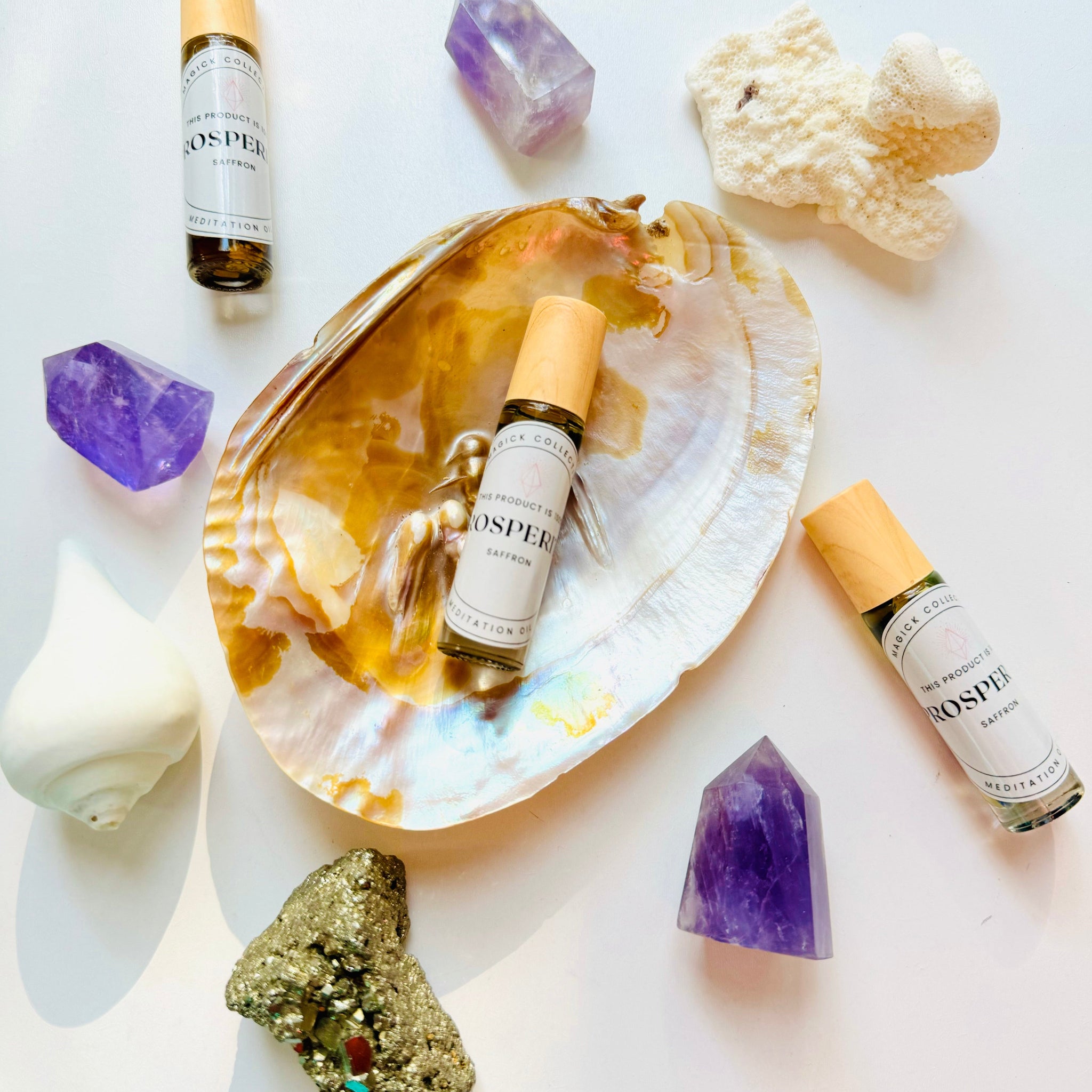 An overhead flat lay showing three "PROSPERity" essential oil roller bottles arranged with several amethyst crystals, a piece of pyrite, and white coral/shells, all centered around a large iridescent seashell.