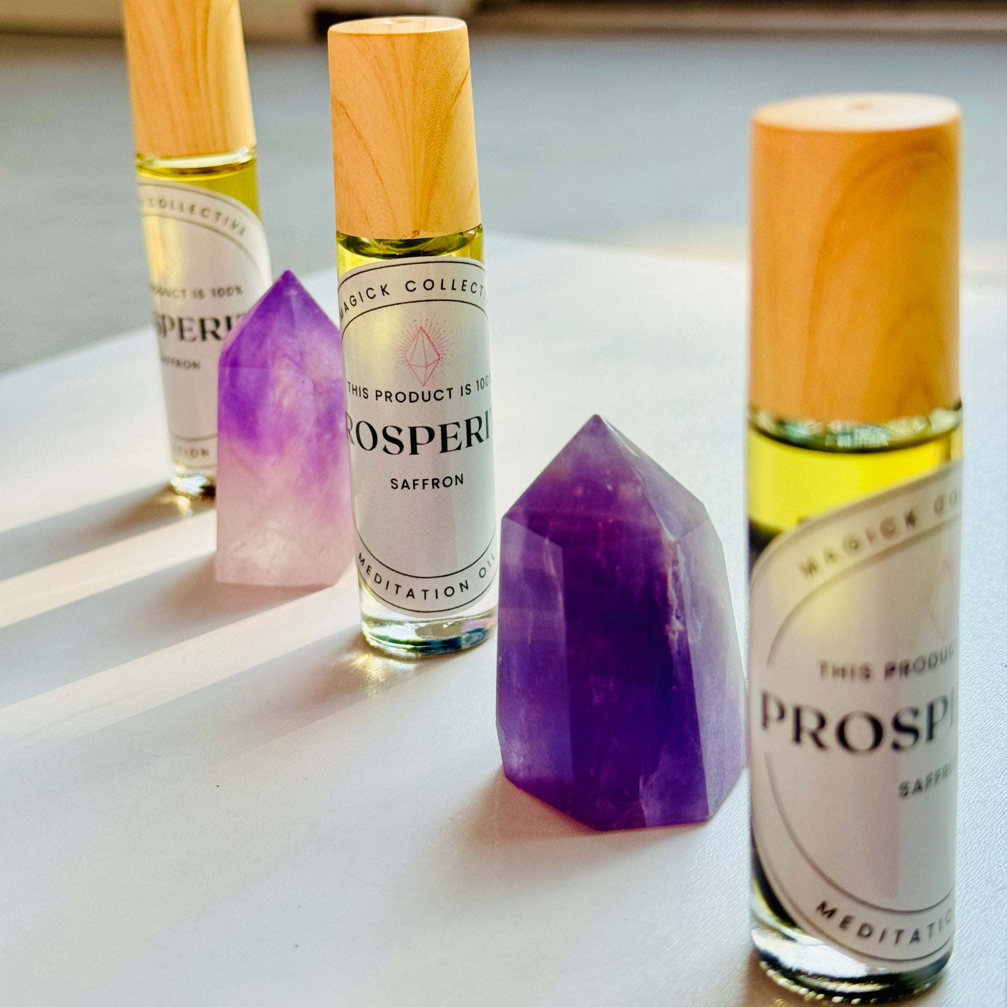 Three small bottles of Prosperity Saffron Perfume Oil with wooden caps, aligned next to each other on a light surface. Each bottle is accompanied by a crystal - yellow, pink, and purple - corresponding to the colors of saffron.