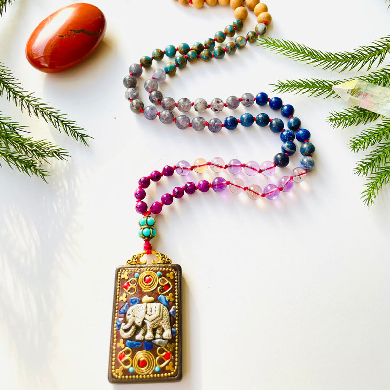 beaded gemstone necklace with elephant pendant on white surface with red gemstone and pine needles