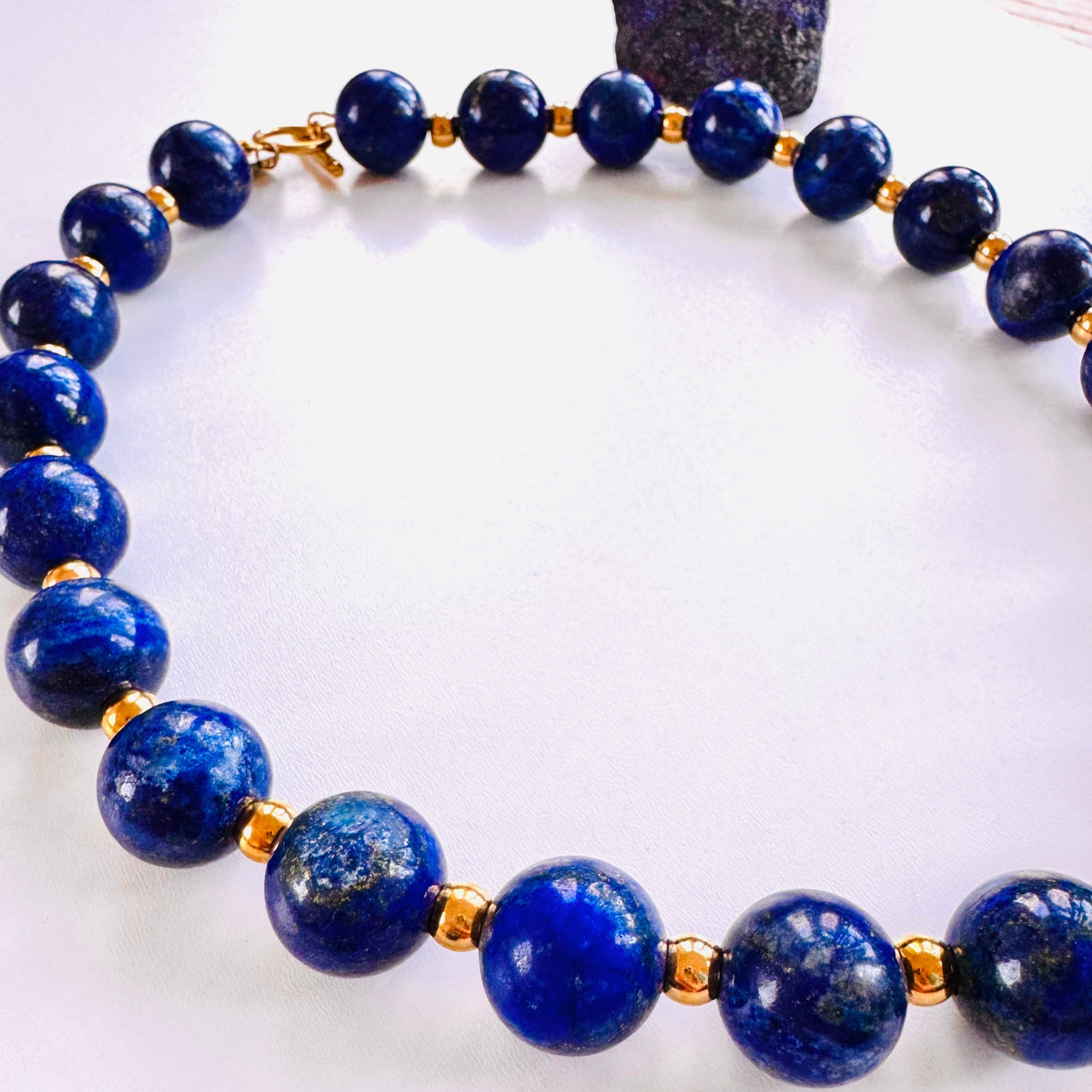 A beaded necklace with blue lapis lazuli and gold-colored accents, displayed against a white background with a large lapis lazuli piece to the side.