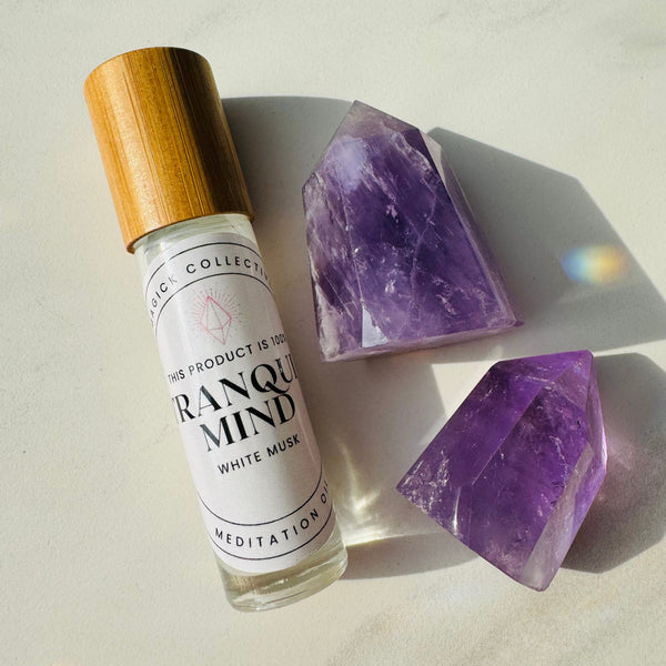 A small bottle of Tranquil Mind Perfume Oil with a wooden cap, labeled with the product name and branding, alongside two pieces of polished purple amethyst gemstones.