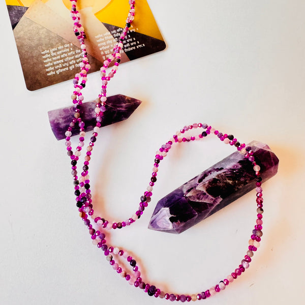 A tantric necklace with a lepidolite gemstone in the center, surrounded by multicolored beads on a silver-colored chain, with a yoga card behind it.