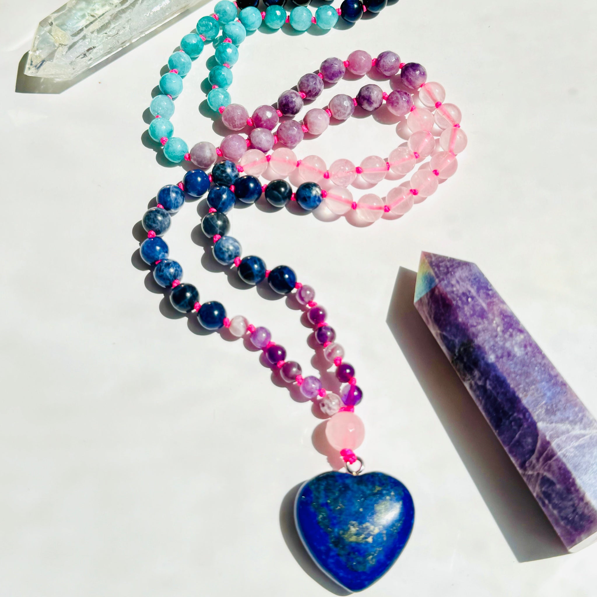 A multicolored beaded mala with a heart-shaped Lapis Lazuli pendant, displayed alongside a crystal and a purple heart-shaped mala.
