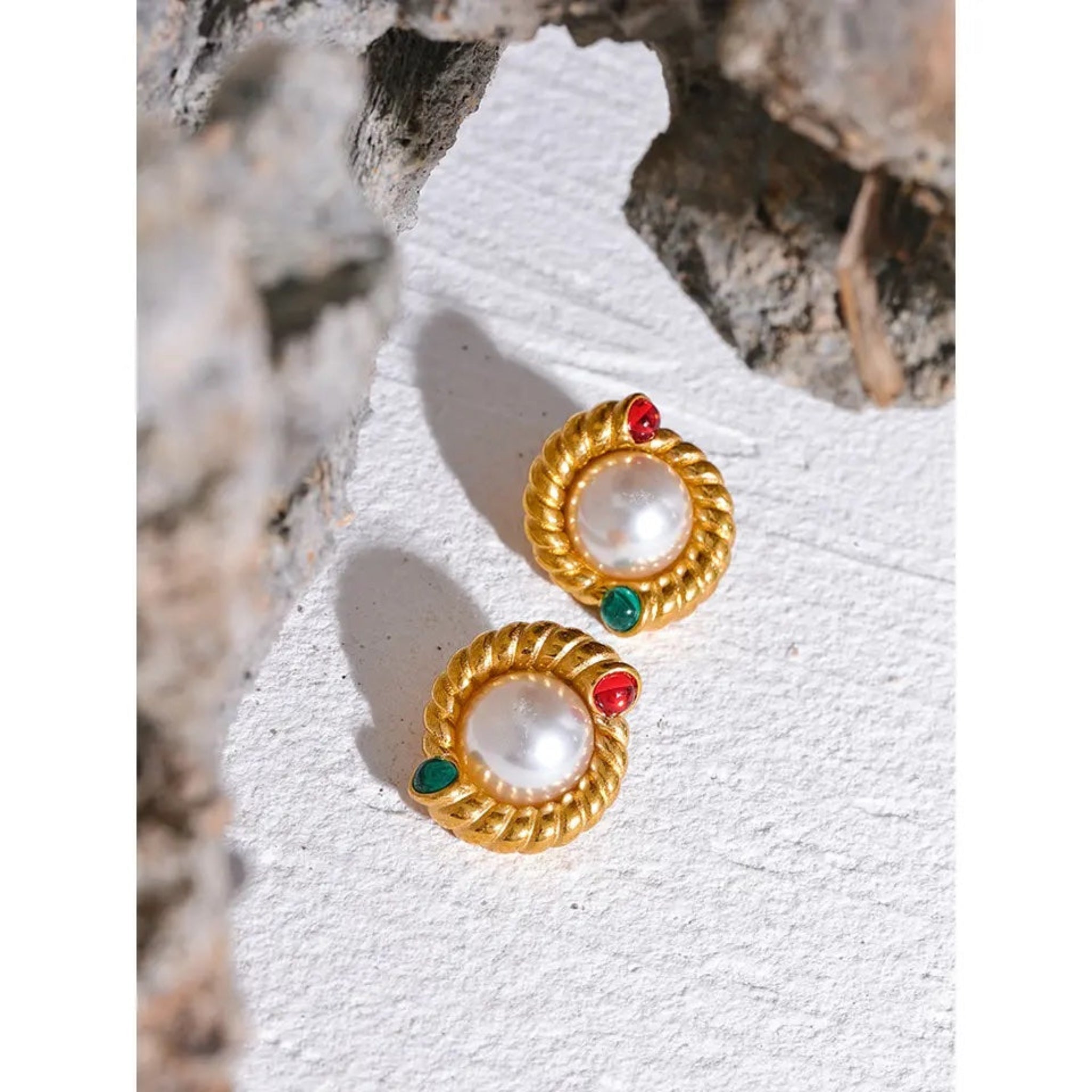 Gold stud earrings with red and green gemstones on a textured white surface.