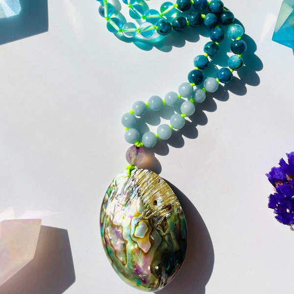 A section of an aqua-colored beaded necklace with a variety of geometric shaped beads and a sea shell pendant, displayed against a white background with other similar items in the background.