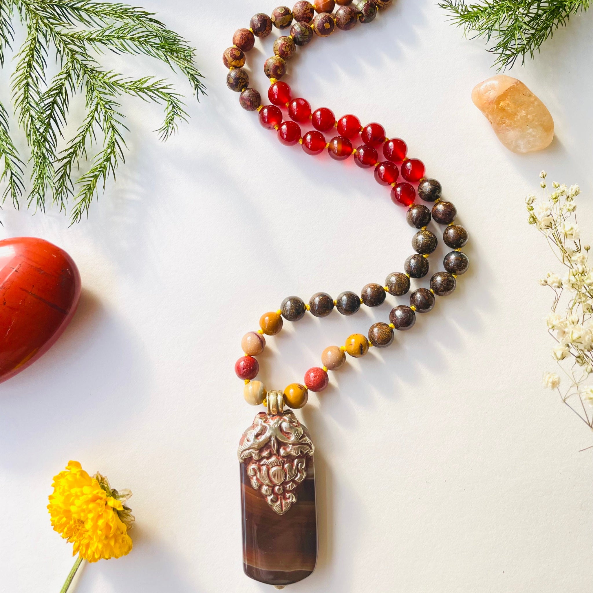 A beaded necklace with a variety of earthy tones and a vintage brass and agate carved pendant, displayed on a white background with natural elements in the background.