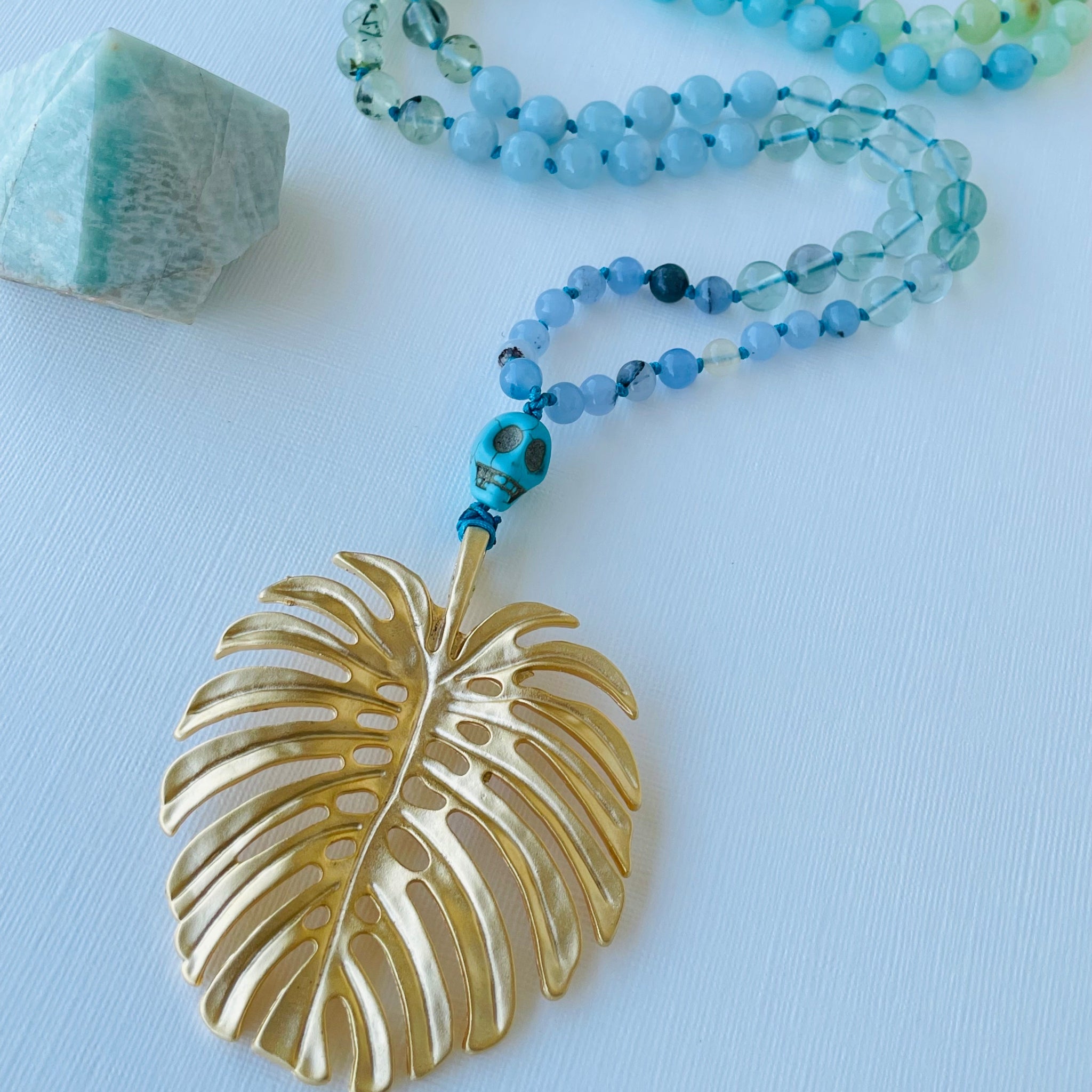 A green and blue beaded necklace with a gold-colored leaf-shaped pendant, displayed against a white background.