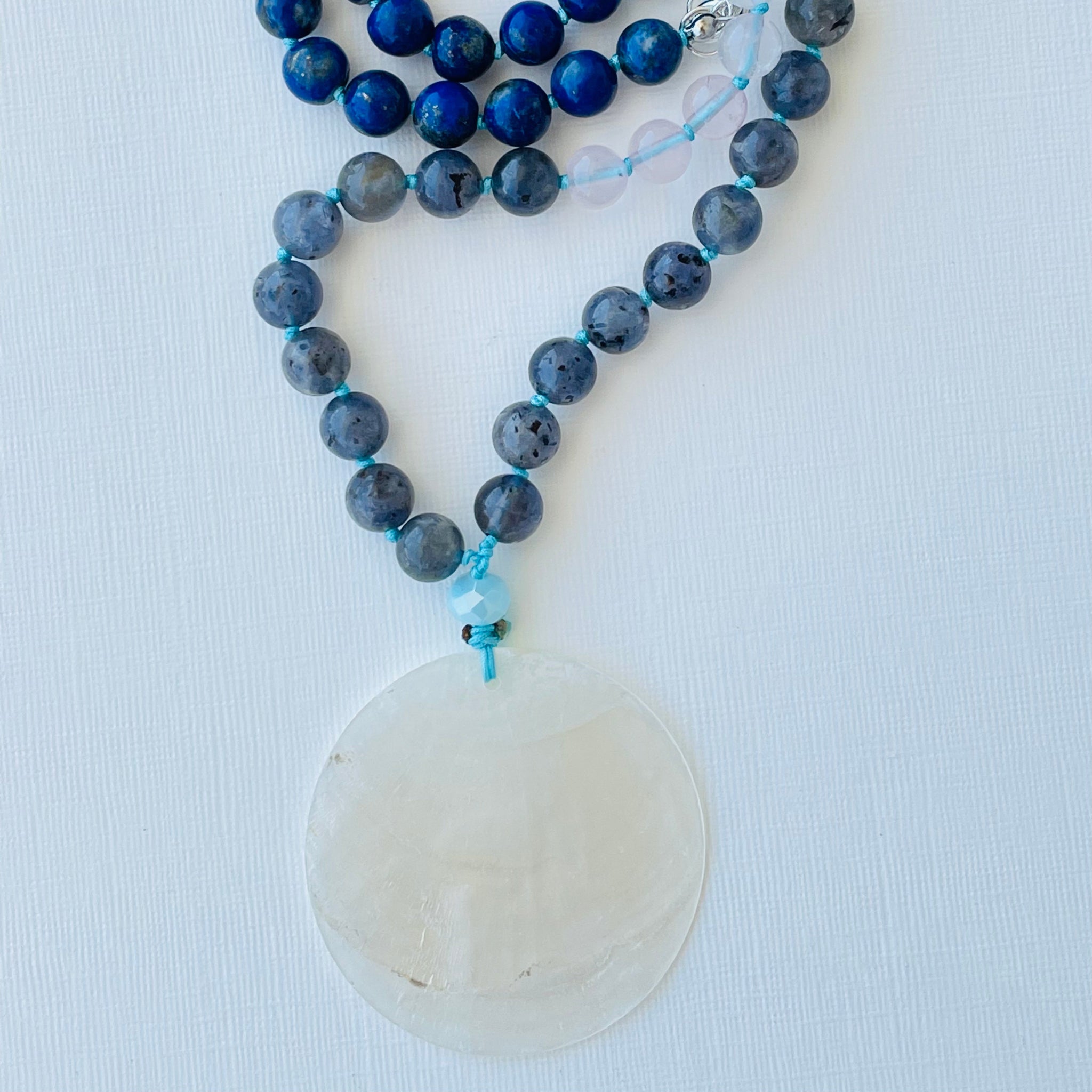 A half mala necklace with a combination of blue and clear beads, a selenite bead, and a nacre pendant, displayed against a white background.