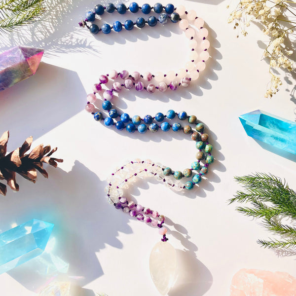 A close-up image of a colorful beaded mala necklace with a variety of natural gemstones, placed on a white surface with decorative items around it, including a crystal and a sprig of rosemary.