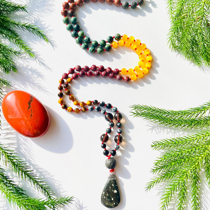 beaded gemstone mala with pine needles and red crystal on white surface