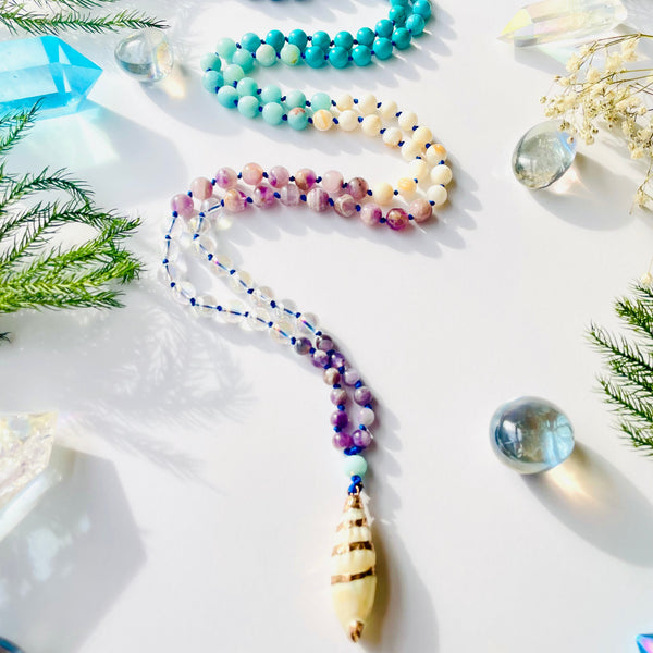 A variety of colorful beads arranged on a white surface, with a question mark-shaped bead in the center, resembling a mala. Some beads are round, while others have a more organic shape, and there are crystal-like elements among them.