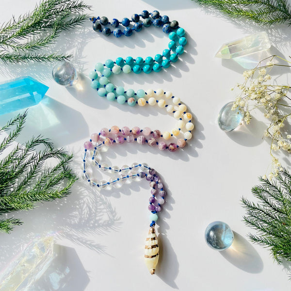 Beaded gemstone necklace with purple, blue and white gemstones on white surface with pine needles and crystals as decor