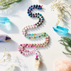 A colorful mala necklace with a mix of gemstone beads and a prominent amethyst pendant, displayed on a white background alongside other decorative items.