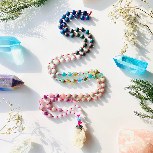 A colorful mala necklace with a mix of gemstone beads and a prominent amethyst pendant, displayed on a white background alongside other decorative items.