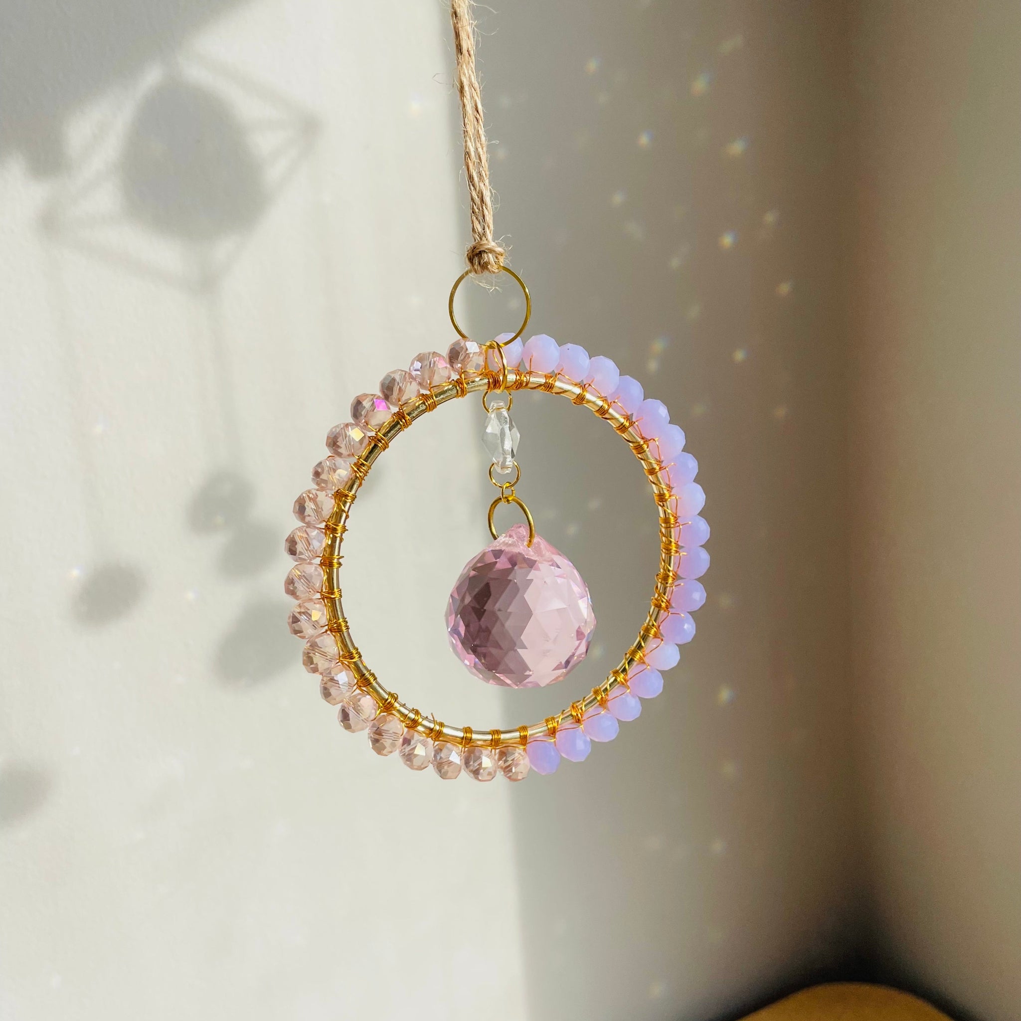 A radiant suncatcher featuring a gold-colored metal frame with blue and pink beads, and a large clear crystal in the center, hanging by a twine against a sunlit background.
