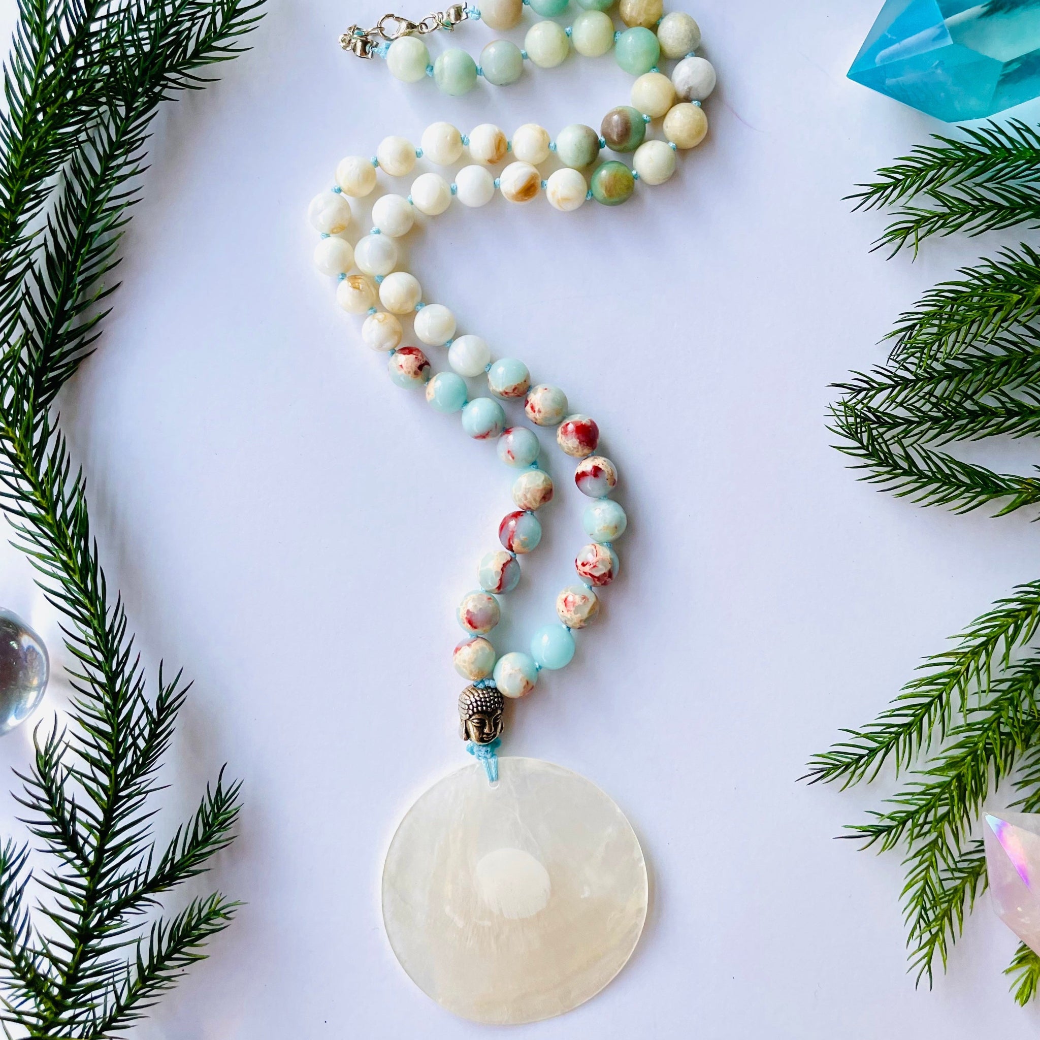 A Mother Of Pearl Half Mala necklace with aquamarine, nacre, and variscite beads, and a nacre pendant, displayed against a white background with greenery in the background.