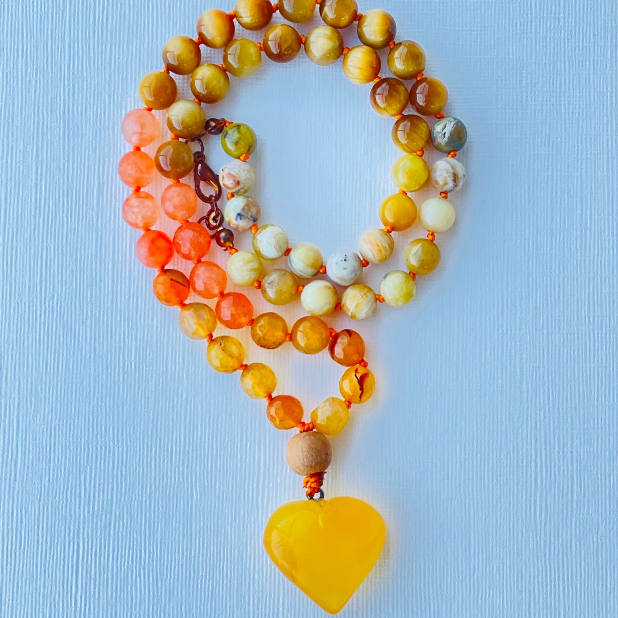 A colorful beaded necklace with a yellow opal, yellow tiger's eye, orange/yellow agate, and amber pendant, displayed on a blue background.