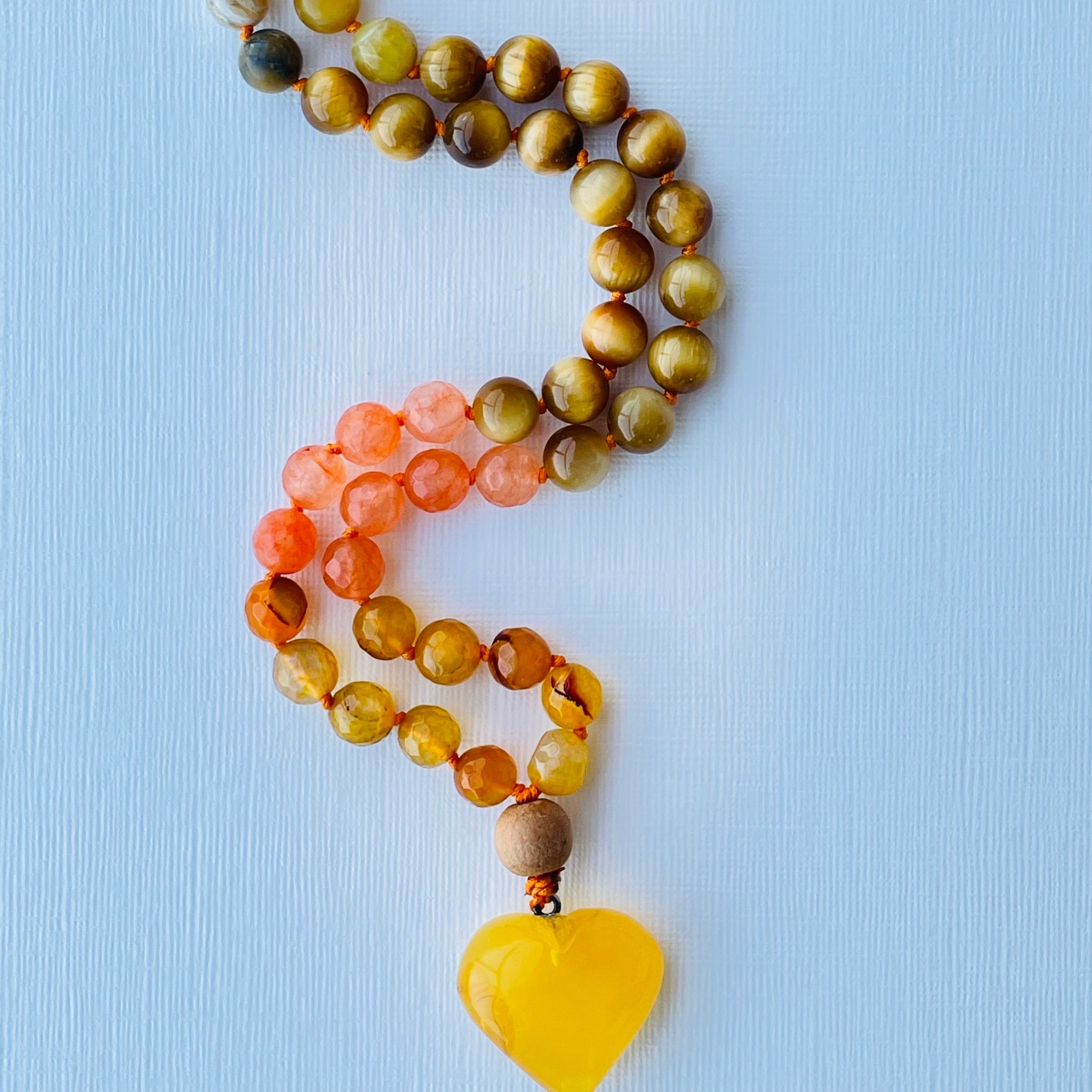 A colorful beaded necklace with a yellow opal, yellow tiger's eye, orange/yellow agate, and amber pendant, displayed on a blue background.
