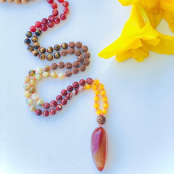 A colorful beaded mala with a red and yellow pendant, displayed on a white surface with sea shell and yellow flowers in the background.