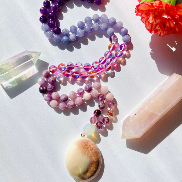 A necklace with purple and pink beaded chains and a seashell pendant, displayed against a white background with floral patterns.
