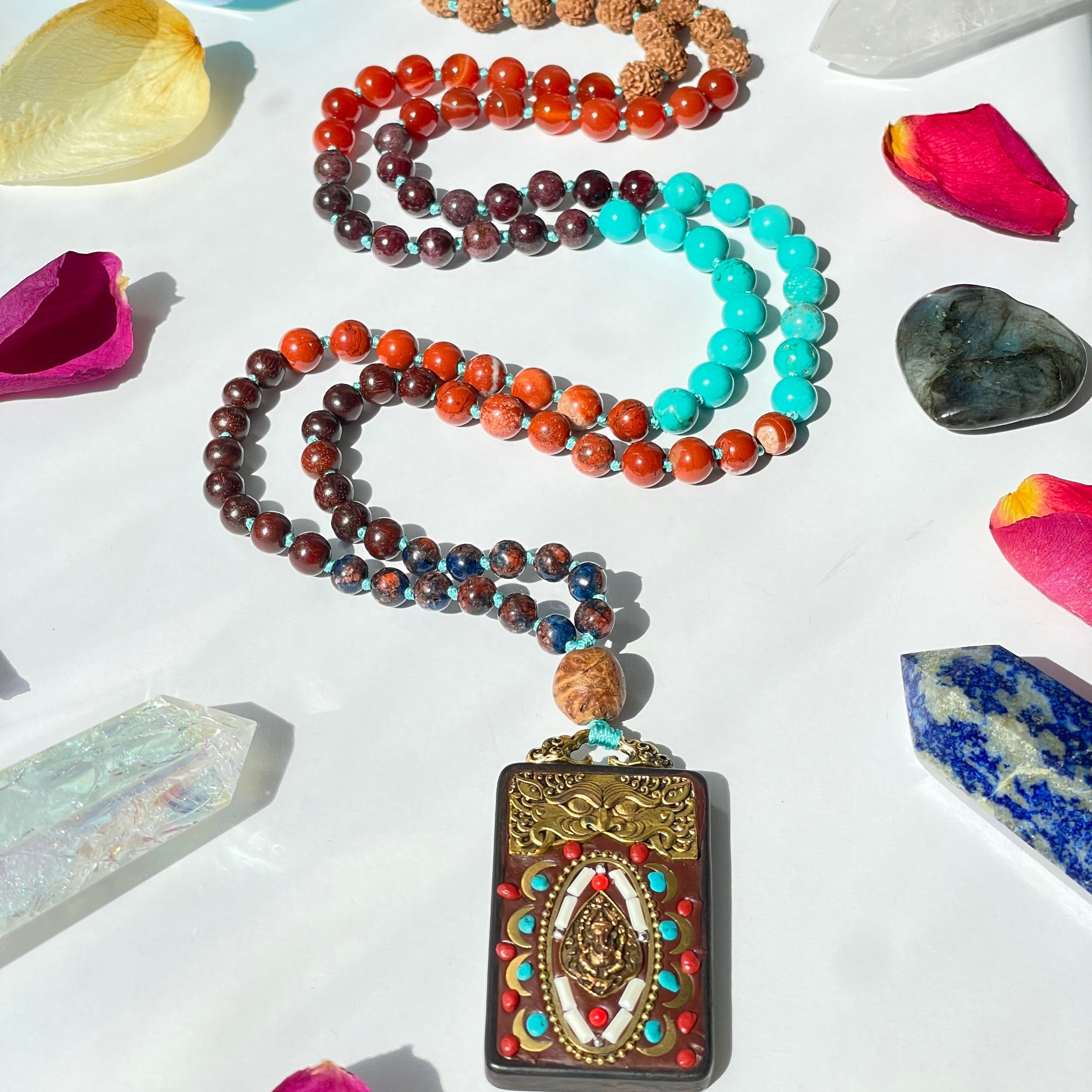 A colorful mala necklace with a mix of beads laid out on a surface with a crystal, a pendant with a deity figure, and petals scattered around.
