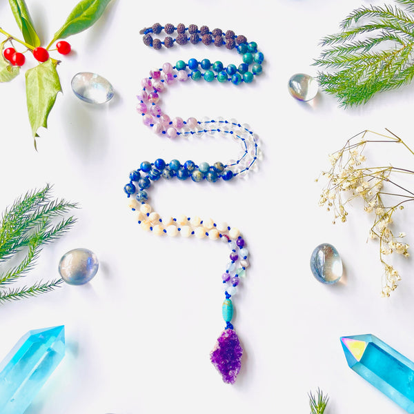 A colorful beaded necklace with a variety of gemstone beads and a purple amethyst pendant, displayed on a white background with decorative crystals and foliage around it.
