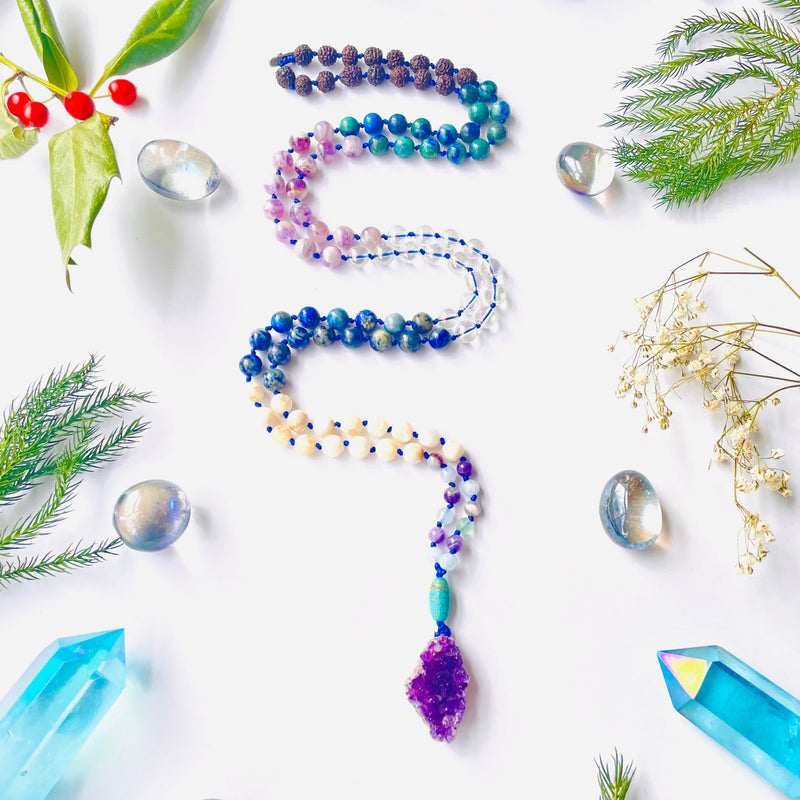 A colorful beaded necklace with a variety of gemstone beads and a purple amethyst pendant, displayed on a white background with decorative crystals and foliage around it.