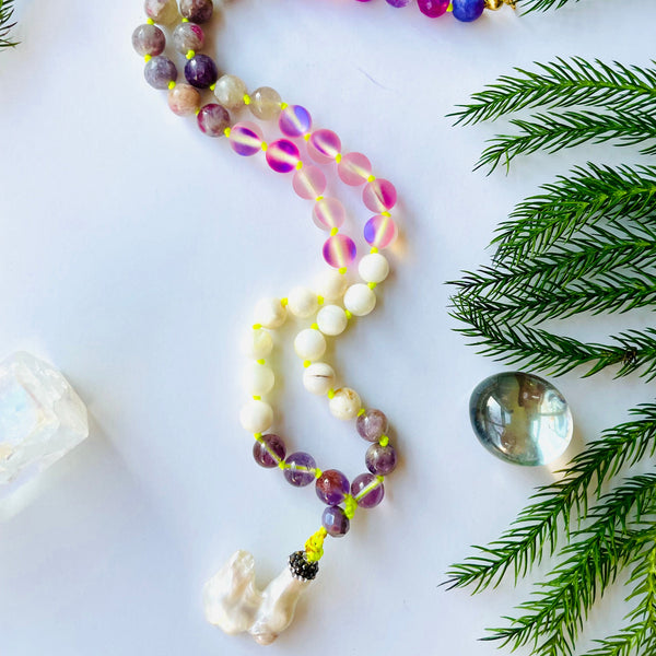 A handcrafted beaded necklace with pink and purple gemstones, featuring a variety of shapes and sizes, displayed on a white background with decorative elements like a pearl pendant and a crystal bead.