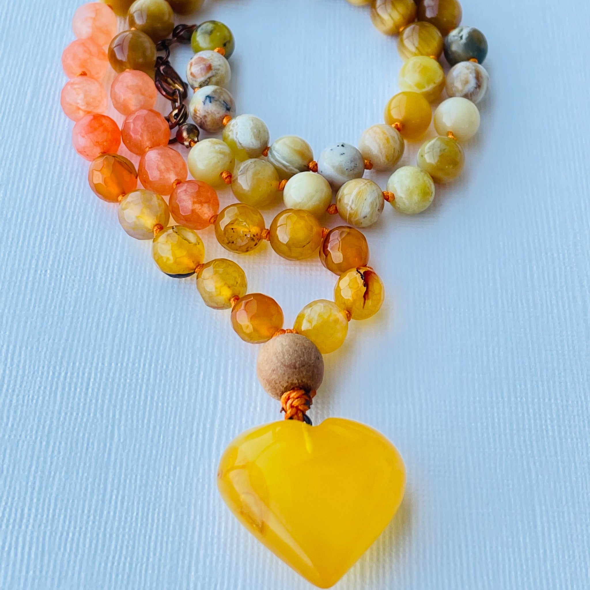 A colorful beaded necklace with a yellow opal, yellow tiger's eye, orange/yellow agate, and amber pendant, displayed on a blue background.